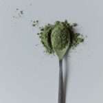 Top view of organic matcha tea powder on a silver spoon against a gray background.